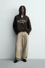 Sudadera de corte boxy fit en tejido de algodón con textura acanalada, color marrón oscuro. Presenta cuello subido con cierre frontal de cremallera y estampado frontal con texto en contraste.