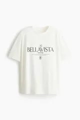 Remera oversized blanca de punto de algodón, con cuello redondo, mangas cortas y estampa tipográfica frontal con la inscripción "The Bellavista".