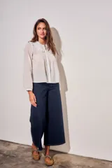 Blusa blanca de algodón peinado con mangas largas, cuello babero bordado y pliegues frontales.