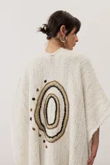 Ruana de punto color beige con detalles geométricos negros y flecos largos tricolores en el borde inferior.