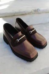 Mocasín de cuero vacuno color bordó con suela track negra de goma. Presenta un diseño de punta cuadrada con un herraje metálico plateado sobre una banda de gamuza en la capellada.