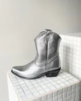 Bota estilo tejana de cuero vacuno en color plateado metalizado, con taco bajo cuadrado y costuras decorativas en la puntera.