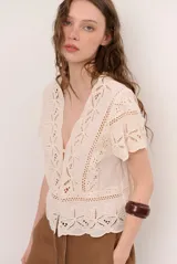 Camisa de broderie color crudo, con escote en V pronunciado, mangas cortas y cierre frontal con botones. Presenta diseños florales calados en toda la prenda y detalles de terminaciones onduladas.