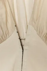 Top beige de manga larga con escote en pico, mangas abullonadas y puños elásticos. Presenta un diseño combinado con tejido fluido en la parte superior y tejido estructurado tipo corsé en la cintura, con cierre frontal de ganchos metálicos.