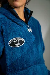 Canguro de textura tipo peluche en color azul, con cierre frontal parcial, cuello alto y bolsillo tipo canguro. Presenta un parche ovalado en el pecho con diseño gráfico.