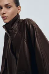 Chaqueta bomber de piel color marrón oscuro, con cuello alto, cierre frontal con cremallera y solapa con botones ocultos. Presenta mangas largas, bolsillos delanteros y un diseño de corte recto.