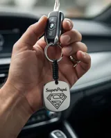 Llavero de acero inoxidable con placa cuadrada grabada con el logo de Superman y la frase Super Papá, unido a una correa trenzada de color negro y argolla metálica circular.