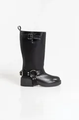 Bota de caña alta estilo biker, confeccionada en cuero sintético negro. Presenta una suela track con taco cuadrado, una hebilla ajustable en la parte superior de la caña y un arnés decorativo en el tobillo con argolla metálica y remaches.