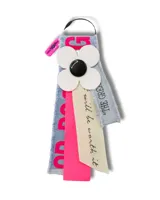 Llavero con base de denim, flor blanca de cuatro pétalos con centro negro, cinta de tela rosa y cinta de tela color crema con texto en negro.