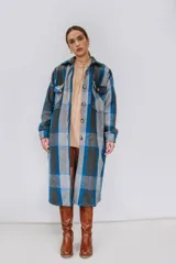 Tapado largo estilo camisa confeccionado en paño italiano, con diseño a cuadros en tonos de azul, gris y negro. Cuenta con cuello clásico, cierre frontal con botones, bolsillos con solapa en el pecho y mangas largas.