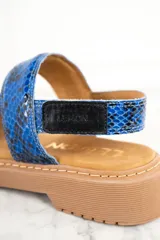 Sandalias con estampado de piel de serpiente en tonos azules, con dos tiras anchas sobre el empeine y una tira alrededor del tobillo. La suela es plana y de color marrón claro.