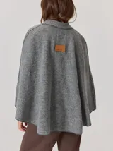 Capa de tejido de punto en color gris melange, con diseño abierto y cierre frontal mediante un botón tipo duffle de madera con detalle en cuero. Presenta bolsillos laterales amplios y bordes con costuras a la vista.
