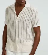 Camisa de manga corta para hombre con cuello solapa y cierre frontal de botones. Presenta un diseño de punto con textura de líneas verticales en color beige y calce regular. Confeccionada en una mezcla de poliéster y algodón sustentable.