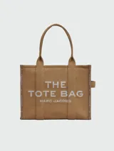Bolso tipo tote de gran tamaño en color beige, confeccionado en lona resistente. Presenta el logo estampado en negro en el frente y detalles de cintas laterales con inscripciones. Cuenta con doble asa superior para llevar al hombro.