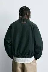 Cazadora de corte relajado color verde oscuro, con cuello de solapa en color marrón a contraste. Presenta cierre frontal con cremallera, bolsillos tipo canguro, puños y ruedo elásticos, y un bordado decorativo en el pecho.