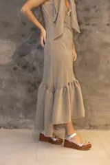 Vestido midi color arena con breteles anchos que se atan en los hombros, escote en V y falda con volado asimétrico en el ruedo. Incluye un aplique decorativo metálico en el centro del pecho.