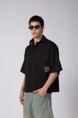 Camisa de manga corta negra 100% algodón, con calce boxy, cuello camisero, cierre frontal con botones y detalle de bordado blanco en la parte inferior.