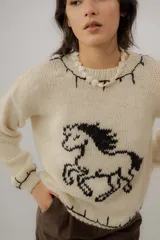 Buzo de punto color crudo, tejido a mano en 100% lana merino, con diseño de caballo en contraste y detalles de pespuntes negros en los bordes.