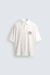 Camisa de corte relajado confeccionada en tejido fluido con mezcla de viscosa, color blanco. Presenta cuello solapa, manga corta, cierre frontal de botones y un bordado a contraste en el pecho.