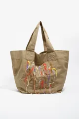 Bolso tipo shopper de color beige, con diseño amplio y doble asa de hombro. Presenta un detalle frontal de bordados coloridos con hilos colgantes que forman un diseño artístico.