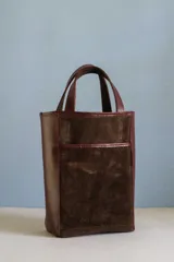 Bolso tipo matera de diseño vertical, confeccionado en una combinación de cuero liso color bordó y gamuza color marrón chocolate. Cuenta con doble asa superior y un bolsillo frontal abierto.