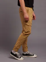 Pantalon tipo cargo color tostado, confeccionado en sarga. Presenta cintura elástica con cordón de ajuste, bolsillos laterales con solapa y puños elásticos en los tobillos.
