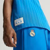 Camiseta de fútbol Adidas del Real Madrid, inspirada en los años 80. Diseño de color azul con finas rayas verticales blancas, cuello en V blanco, mangas cortas con las tres tiras características de la marca y escudo retro del club bordado en el pecho.
