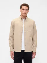 Camisa de estilo Oxford en color beige, con cuello abotonado, cierre frontal de botones, mangas largas con puños abotonados y un bolsillo en el pecho con el logo de un oso bordado.