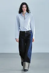 Jeans de tiro bajo y pernera ancha, confeccionados en denim azul oscuro con pespuntes en contraste. Cuentan con un diseño de cinco bolsillos, cierre frontal con cremallera y botón metálico, y destacan por su amplio doblando en el bajo que deja ver el revés de la tela con detalles de costura selvedge.