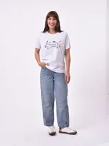 Remera de manga corta color beige claro con cuello redondo. Presenta un estampado gráfico central con un plato de pasta rodeado por un borde azul y blanco, sobre un fondo de franjas verticales rojas y blancas. El estampado incluye texto en cursiva en tonos turquesa y rojo.