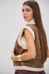 Chaleco de estilo aviador en color camel, confeccionado en material efecto cuero con interior y bordes de corderito sintético color crema. Presenta cierre frontal, cuello solapa y detalles de hebillas metálicas en los laterales y el cuello.