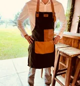 Delantal de cocina color verde musgo con detalles en cuero color suela. Tiene tiras de cuero ajustables al cuello y a la cintura, y dos bolsillos frontales de cuero. Ideal para asados y cocina en general.