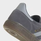 Championes Adidas Handball Spezial de gamuza color gris con las icónicas tres tiras laterales en un tono más claro y suela de caucho natural. Presenta el logo de la marca y el nombre del modelo en dorado en el lateral.