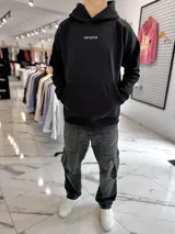 Canguro oversize negro con capucha y bolsillo tipo canguro. Presenta un estampado gráfico en la espalda con la inscripción "TOP STYLE", un rectángulo con efecto marmolado en verde y negro con la palabra "WORLDWIDE", y texto descriptivo adicional con un código de barras.