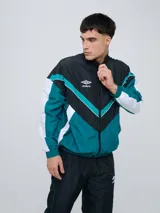 Campera deportiva Umbro modelo Beat, confeccionada en nylon ligero con diseño de bloques de color en blanco, azul marino y verde azulado. Presenta cierre frontal, cuello alto y elásticos en puños y cintura.