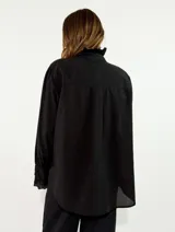 Camisa de algodón color blanco, con cuello redondo y detalle de volados en el cuello y puños. Presenta mangas largas con puños elásticos y un diseño de corte holgado con ruedo asimétrico, más largo en la parte trasera.