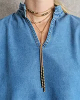 Collar estilo corbatín confeccionado en tiento de cuero color suela, con detalle de cuentas metálicas plateadas distribuidas a lo largo de las tiras.