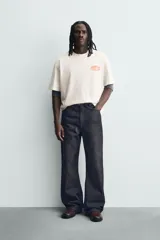 Camiseta de corte relajado color crudo, con cuello redondo y manga corta. Presenta un estampado gráfico pequeño en el pecho y un diseño grande en la espalda relacionado con el artista Post Malone.