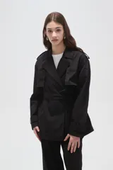 Trinchera de corte corto y diseño clásico, confeccionada en poliéster color negro. Presenta solapas amplias, cierre frontal cruzado con lazo ajustable en la cintura, charreteras en los hombros y puños con trabillas.