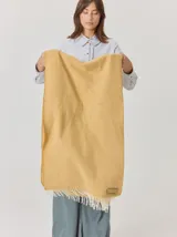 Bufanda de gran tamaño en color mostaza con diseño de tejido en espiga y flecos en los extremos. Incluye una etiqueta metálica decorativa en una de las esquinas.