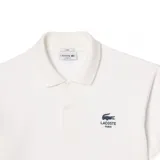 Chomba Lacoste de piqué color blanco, de manga corta, con cuello camisero y cierre frontal de dos botones. Cuenta con el logotipo de la marca bordado en el pecho junto con el texto "Lacoste Paris".