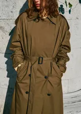 Trench coat de algodón en color verde oliva, con diseño largo y oversize. Presenta cuello camisero de pana, cierre de doble botonadura, cinturón con hebilla, trabillas en hombros y puños ajustables.