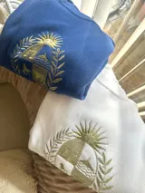 Buzo de tejido de punto color azul, con cuello redondo y diseño holgado. Cuenta con un gran bordado dorado en el pecho que representa el escudo patrio rodeado de laureles.