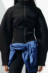 Chaqueta negra de cuello subido con cierre frontal de cremallera termosellada, manga larga con puños anchos y cremalleras laterales en los puños.