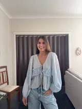 Camisa de tela liviana color celeste, con escote en V, mangas largas abullonadas con puños elásticos y detalle de volado fruncido en el frente con cierre de cordones ajustables.