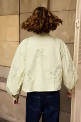 Camisa de algodón color blanco con cuello mao y cierre frontal de botones. Presenta un diseño de bordados florales en color negro distribuidos en el frente y las mangas. Posee mangas largas abullonadas con puños abotonados y un corte recto ligeramente cropped.
