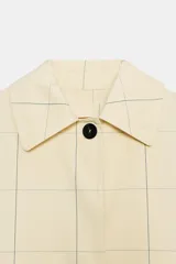 Trench de algodón color amarillo claro con patrón a cuadros sutiles de líneas oscuras. Tiene cuello con solapa, mangas largas, bolsillos delanteros y cierre frontal con botones ocultos bajo solapa.