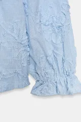 Blusa de escote pico y manga larga con puño elástico y volante. Presenta un diseño de rayas verticales finas en azul claro y blanco, con flores en relieve.