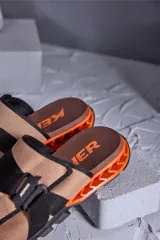 Chinelas Kenner modelo Rakka Slide Pro, con capellada de tela color beige y ajuste mediante hebilla técnica negra. Presenta una base ergonómica marrón con estructura lateral geométrica en color naranja y suela negra.