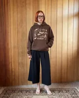 Canguro oversize color marrón chocolate, con capucha y bolsillo frontal tipo canguro. Presenta un estampado gráfico en color crema en el pecho con diversos dibujos y la inscripción 'Más barrio que moda' y 'Kabor'.